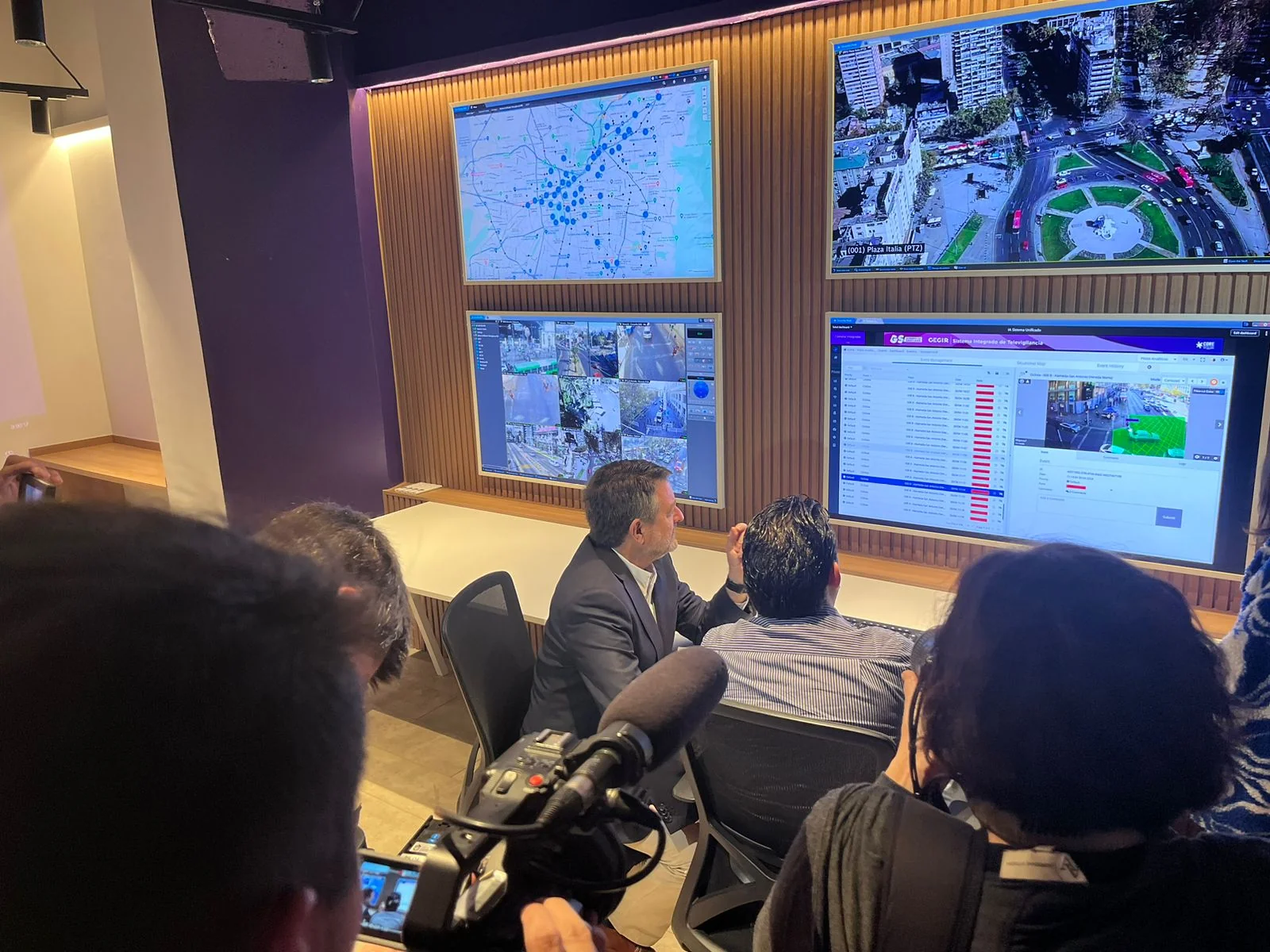 The Governor of Santiago seated at the Sala CEGIR operator desk being briefed by technical staff while press record from behind.